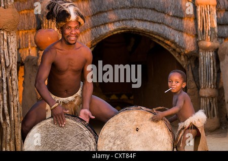 Zulu uomo in costume tradizionale a suonare la batteria, set cinematografico di Shakazulu, Shakaland, KwaZulu-Natal, Sud Africa e Africa Foto Stock