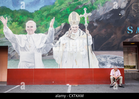 Uomo seduto su uno sgabello sotto un murale del Papa a Lourdes, Hautes Pirenei, Francia Foto Stock