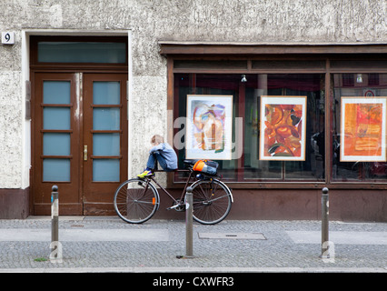 Un giovane ragazzo bilanciamento sulla traversa di una bicicletta di fronte a un negozio di arte a Berlino Foto Stock