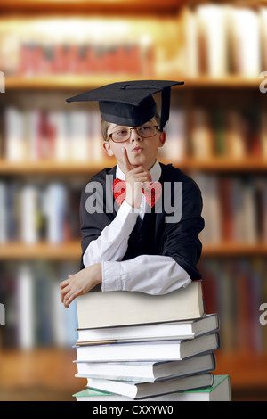 Riflessivo cercando schoolboy indossando un cappello di graduazione e appoggiato su di un pile di libri in una libreria Foto Stock