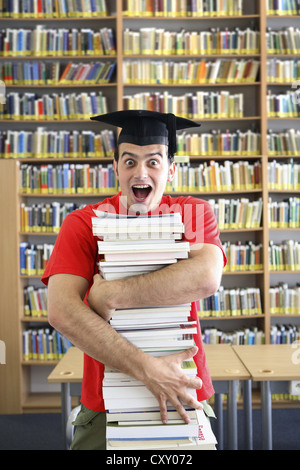 Studente indossando un cappello di graduazione e titolari di una pila di libri in una biblioteca universitaria Foto Stock