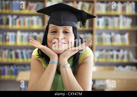 Sorridente studentessa indossando un cappello di laurea in una biblioteca universitaria Foto Stock