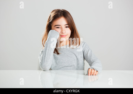 Bella ragazza seduta dietro una scrivania e sorridente, contro uno sfondo grigio Foto Stock