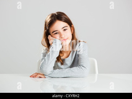 Bella ragazza seduta dietro una scrivania con un espressione distratto, contro uno sfondo grigio Foto Stock