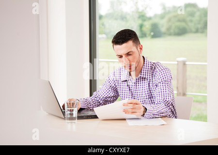 Giovane uomo seduto alla sua scrivania e lavorare attraverso i documenti Foto Stock