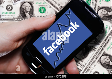 Mano che tiene uno smartphone con un logo di Facebook di fronte le fatture del dollaro, immagine simbolica per la IPO di Facebook Foto Stock
