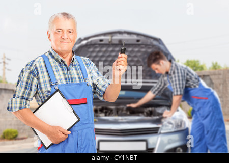 Coppia meccanico in uniforme tenendo premuto un tasto auto e un altro meccanico di eseguire una verifica automobile in background Foto Stock