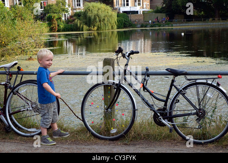 Un bambino gioca con un grazioso laghetto su Hampstead Heath. Egli è circondato da biciclette e gioca con un bastone. Foto Stock