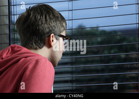 Giovane uomo che indossa gli occhiali, guardando attraverso una finestra cieca. Foto Stock