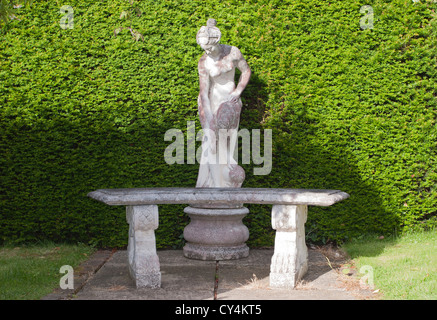 Una statua di pietra e sedersi al banco in un paese di lingua inglese giardino anteriore di una siepe verde Foto Stock