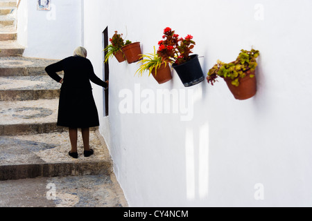 Vecchia vedova spagnola vestita di nero per la sua strada verso il lastricato in ciottoli scale di una corsia dipinte di bianco decorato con colorati vasi. Foto Stock