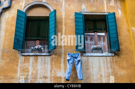 Sbiadito e colorate di giallo ocra-parete, finestre con persiane blu e un paio di blue jeans denim appendere fuori ad asciugare in Venezia Foto Stock