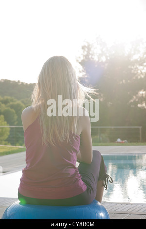 Donna seduta su sfera fitness a bordo piscina, vista posteriore Foto Stock