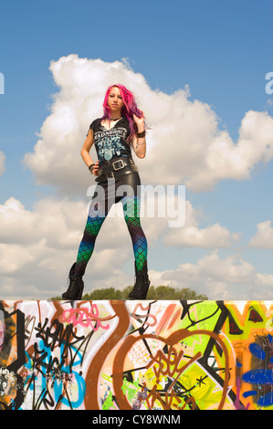 Giovani donne/ ragazza con i capelli rosa e nero mini dress seduto sul muro, contro il cielo blu con la nuvola bianca Foto Stock