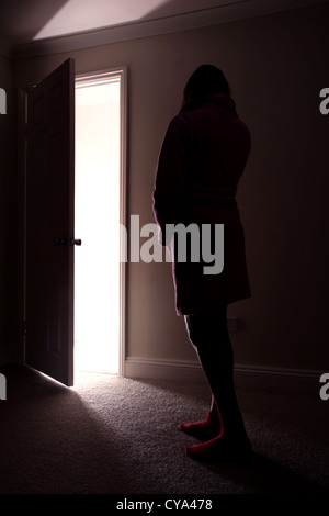 Vista posteriore di una ragazza adolescente in piedi in una stanza buia a guardare verso una luce che risplende attraverso una porta aperta. Foto Stock