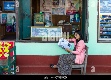 Myanmar, una donna leggendo il giornale davanti a un negozio di orologi nel Hleku street market, lungo la strada che da Yangon a Bago. Foto Stock