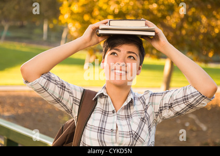 Ritratto di una bella gara di misto femmina giovane azienda libri sul suo capo all'aperto presso il parco in un pomeriggio soleggiato. Foto Stock