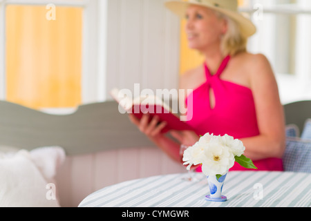 Signora legge un libro sulla veranda, reposeful atmosfera, formato verticale, stretta di concentrarsi sul fiore, signora guarda al di fuori Foto Stock