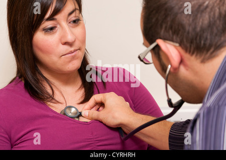 GP ambulatorio medico paziente consultazione UK. Dottore in ascolto al cuore del paziente utilizzando uno stetoscopio. Foto Stock