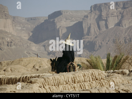 Donna che indossa un alto cono Hat e cavalcando un asino in terra rocciosa, Hadramaut, Yemen Foto Stock