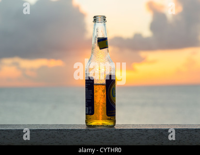 Di ghiaccio freddo Carib bottiglia di birra sul tavolo di fronte tramonto sull'oceano Foto Stock