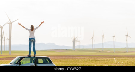 Giovane donna in piedi sulla parte superiore della sua vettura con le mani alzate. Ragazza in Giallo autunno campo con turbine eoliche in background. Foto Stock