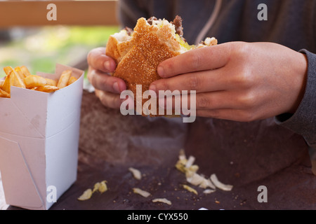 Un close-up di una donna di mani in parte mangiato hamburger con un ordine di patatine fritte. Foto Stock