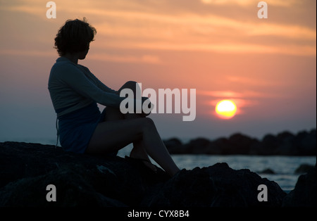 Una giovane donna di godersi il tramonto seduti sulle rocce in una spiaggia. Foto Stock