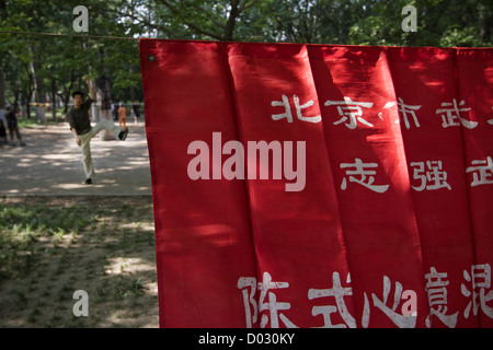 Un uomo che fa una serie di esercizi del mattino dietro un banner rosso con scrittura cinese su di esso in un parco a Pechino in Cina. Foto Stock