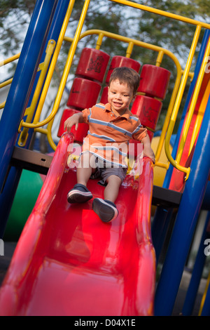 Un bambino di 2 anni boy gioca allegramente su una slitta rossa in un parco giochi all'aperto Foto Stock