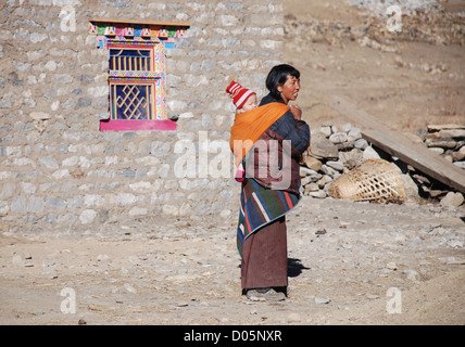 Una donna che indossa tradizionale abito tibetano e portante un bimbo piccolo sorge al di fuori di una casa nella regione dolpo del Nepal Foto Stock