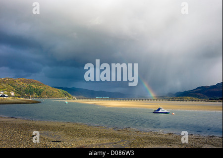 Locale dei pendolari treno passa oltre Barmouth Bridge estuario, sotto un arcobaleno, storm su montagne, barche ormeggiate in primo piano sun Foto Stock