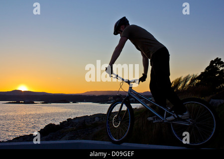 Un trial bike rider saldi su una parete al tramonto con un paesaggio marino e basse colline in background - Nessuna vendita su Alamy o ovunque Foto Stock