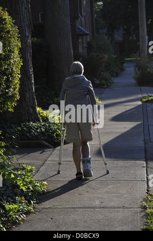 Una donna con un piede destro ferito cammina con cautela su due stampelle, all'aperto, su un marciapiede, Ontario, Canada. Verticale. Spazio di copia. Rihab. Foto Stock