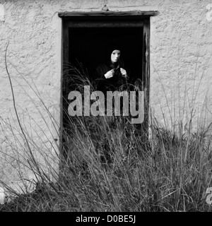 Figura mascherata dalla rottura di porta della casa abbandonata oscurata da piante sovradimensionate. Foto Stock