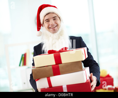 Immagine di happy businessman in Santa Cappello e barba holding stak di doni e guardando la telecamera in ufficio Foto Stock