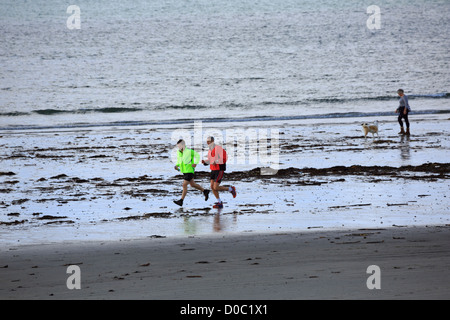 Corsa mattutina, Mounts Bay, Cornwall. Due uomini in esecuzione sulla spiaggia vicino a Marazion mentre una donna che gioca con il suo cane vicino all'acqua. Foto Stock