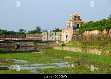 Imperial City Moat Bridge Stone Arch Brick Gate Tower Hue Vietnam / HUE, Vietnam - Un ponte ad arco in pietra attraversa il fossato verso una torre fortificata in mattoni presso la città imperiale. I bastioni in muratura inclinati e i corsi d'acqua difensivi riflettono la disposizione della fortezza in stile Vauban, la prima del suo genere nel sud-est asiatico, che l'imperatore Gia Long incorporò a partire dal 1803 per proteggere la nuova capitale della dinastia Nguyen. Il progetto ha deliberatamente fuso l'ingegneria militare europea con un piano sonoro cosmologico orientato intorno al fiume Perfume e alle colline circostanti. Foto Stock