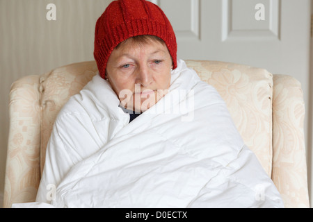 Senior donna sensazione di freddo premuto e da solo indossando un cappello e avvolta in un piumone cercando di mantenere caldo e accogliente a casa in inverno. Inghilterra Regno Unito Gran Bretagna Foto Stock