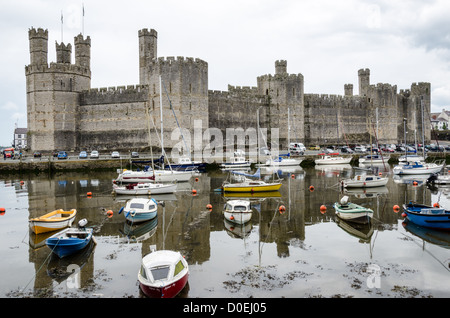 CAERNARFON, Galles — Una vista del castello dall'altra parte del fiume, con barche ormeggiate in primo piano, al castello di Caernarfon nel Galles nordoccidentale. Un castello originariamente sorgeva sul sito risalente alla fine dell'XI secolo, ma alla fine del XIII secolo re Edoardo i commissionò una nuova struttura che si erge fino ad oggi. Ha torri caratteristiche ed è uno dei castelli meglio conservati della serie commissionata da Edoardo I. Foto Stock