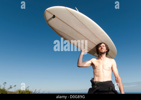 Surfista maschio in piedi con longboard sulla sommità della testa con muta verso il basso per la sua vita, Raglan Nuova Zelanda. Foto Stock