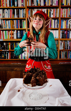 Elf personaggio vestito di rosso e verde di Natale vestito a mangiare una torta al cioccolato in una libreria o biblioteca. Foto Stock