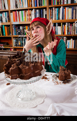 Elf personaggio vestito di rosso e verde di Natale vestito a mangiare una torta al cioccolato in una libreria o biblioteca. Foto Stock