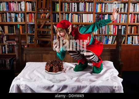 Elf personaggio vestito di rosso e verde di Natale vestito a mangiare una torta al cioccolato in una libreria o biblioteca. Foto Stock