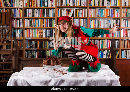 Elf personaggio vestito di rosso e verde di Natale vestito a mangiare una torta al cioccolato in una libreria o biblioteca. Foto Stock