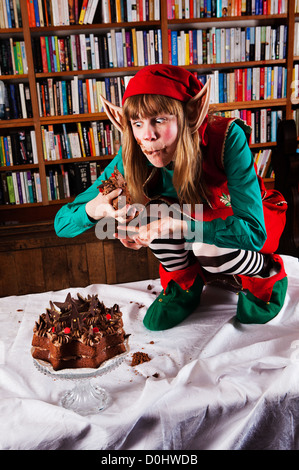 Elf personaggio vestito di rosso e verde di Natale vestito a mangiare una torta al cioccolato in una libreria o biblioteca. Foto Stock