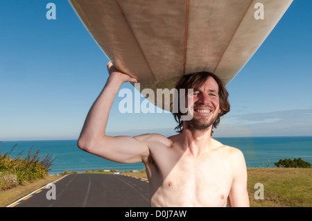 Surfista maschio in piedi con longboard sulla sommità della testa con muta verso il basso per la sua vita, Raglan Nuova Zelanda. Foto Stock