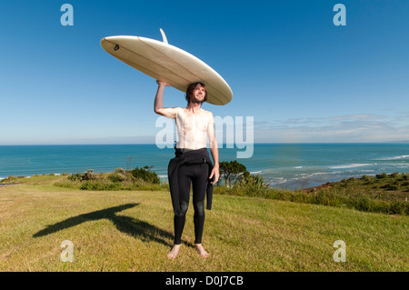 Surfista maschio in piedi con longboard sulla sommità della testa con muta verso il basso per la sua vita, Raglan Nuova Zelanda. Foto Stock