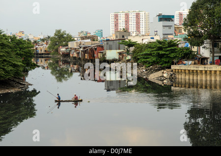 HANOI, Vietnam - due uomini in una piccola canoa di legno usano una rete per pescare nelle acque tranquille di un affluente del fiume Saigon nella città di ho chi Minh, Hanoi, con case improvvisate sul lungomare e alti altipiani sullo sfondo. Foto Stock