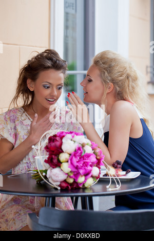 Due belle ragazze in estate abiti gossip al tavolo del bar con un mazzo di fiori e un dessert su di esso Foto Stock